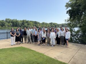 Participants of the Busting Water Myths workshop pose for a group photo on a riverside overlook along the Black Warrior River. The group stands on a paved terrace beside a black metal railing, with the calm river, tree-lined shoreline, and clear blue sky forming the backdrop. A grassy lawn is visible in the foreground, highlighting the outdoor setting and connection to the watershed-focused workshop.