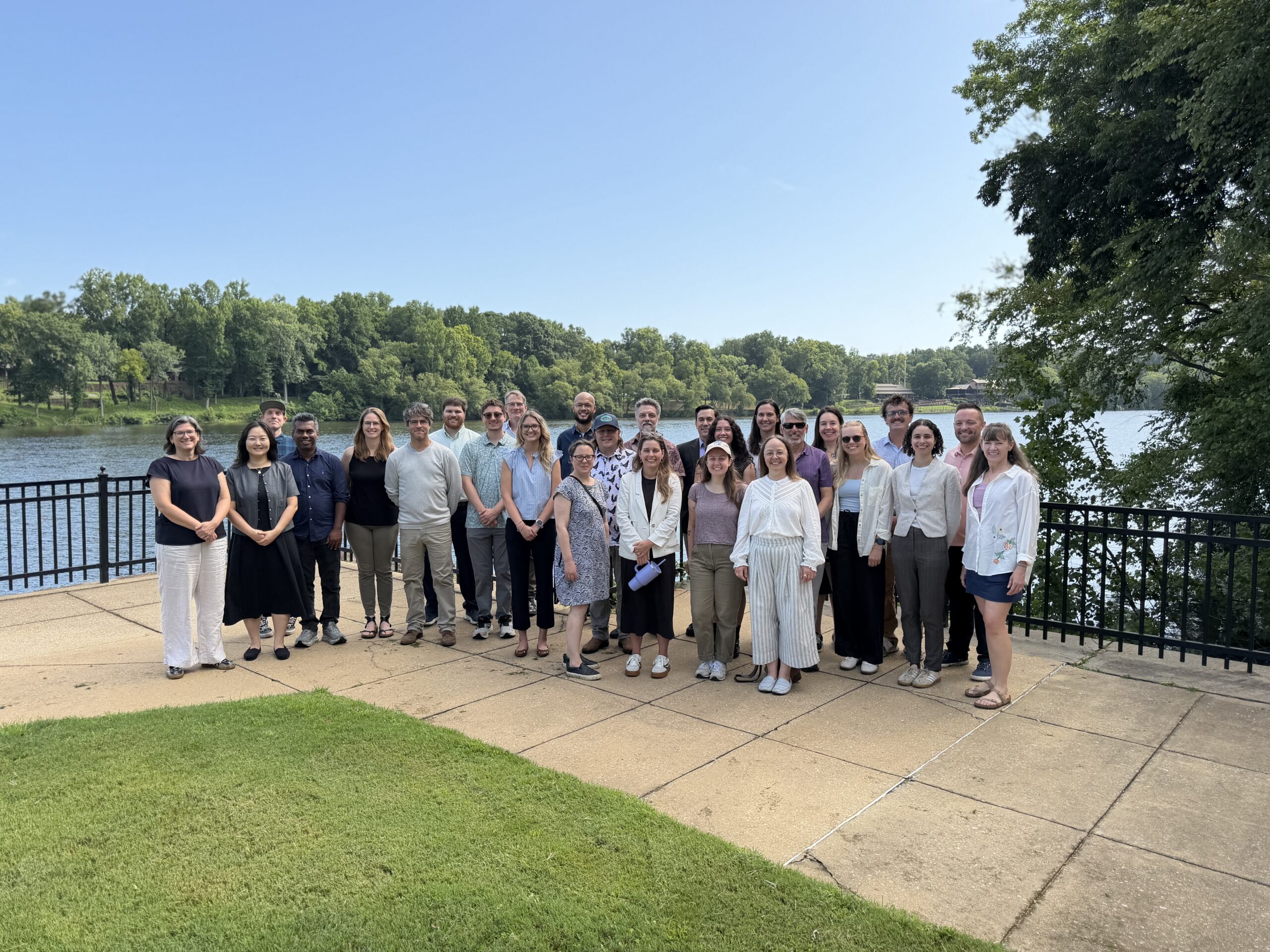Participants of the Busting Water Myths workshop pose for a group photo on a riverside overlook along the Black Warrior River. The group stands on a paved terrace beside a black metal railing, with the calm river, tree-lined shoreline, and clear blue sky forming the backdrop. A grassy lawn is visible in the foreground, highlighting the outdoor setting and connection to the watershed-focused workshop.