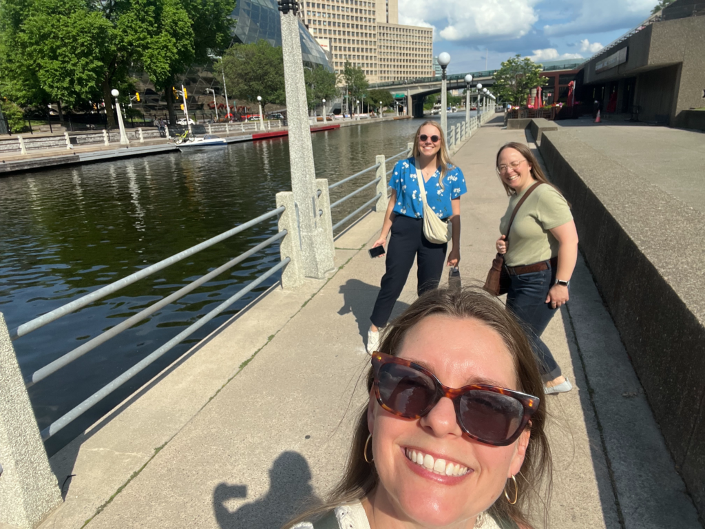 Three women stand beside a scenic urban river with a safety railing and large buildings in the background. The women in the photo are Penny Beames, Dr. Penelope Mitchell, and Dr. Caily Schwartz